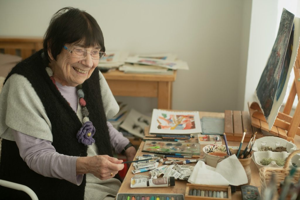 A older person smiling in front of an artist's easel, with lots of paints