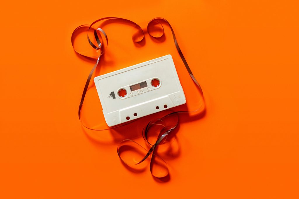 A photograph of a white cassette tape being re-wound against a block bright orange-coloured backdrop