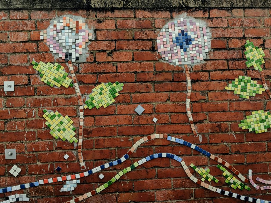 Colourful mosaic depicting two flowers with stems and leaves, against a red brick wall