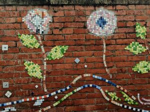 Colourful mosaic depicting two flowers with stems and leaves, against a red brick wall