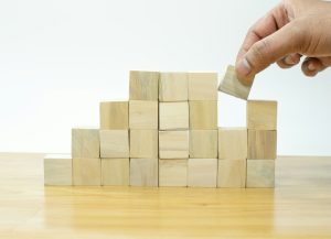 Photograph of a hand placing a small wooden building block onto a stack of wooden building blocks
