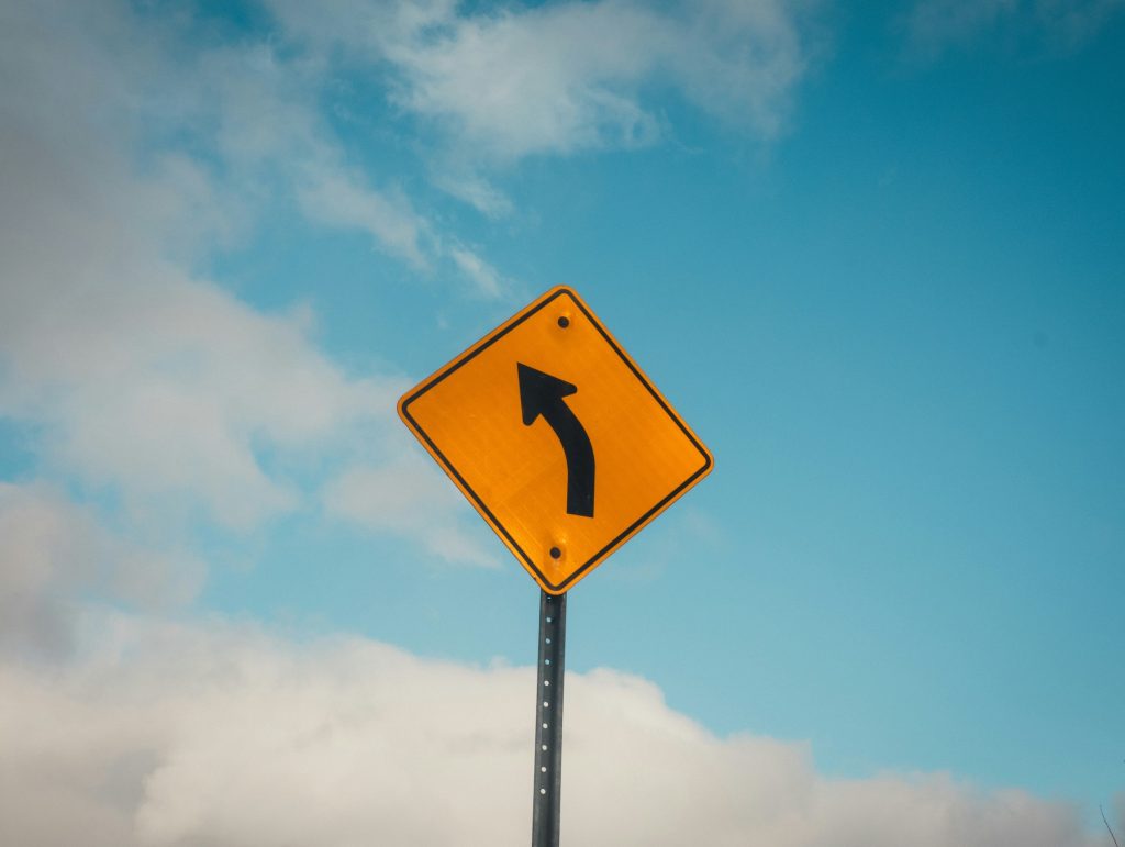 Photograph of a yellow road sign with a black arrow pointing left, against a cloudy blue sky