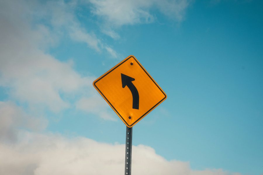 Photograph of a yellow road sign with a black arrow pointing left, against a cloudy blue sky